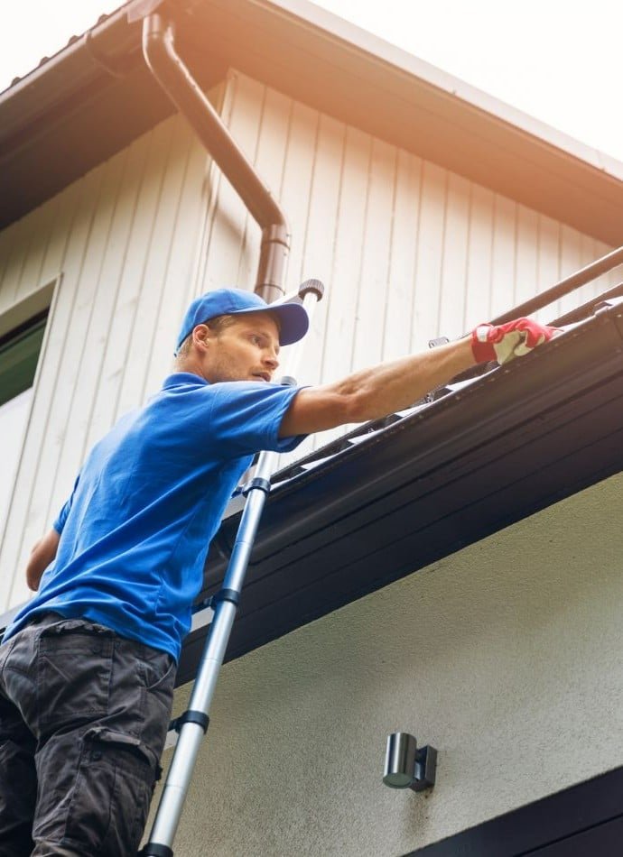 Man on ladder cleaning gutters of a house. He's wearing a blue shirt and hat, with work gloves, reaching into the dark metal gutter to remove debris. Gutter cleaning is important for home maintenance.