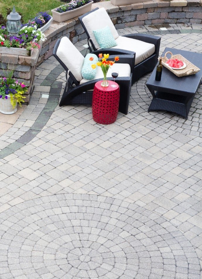Outdoor patio featuring concrete pavers in a circular design, with lounge chairs, a red accent table, and flowers, showcasing a beautiful backyard space.