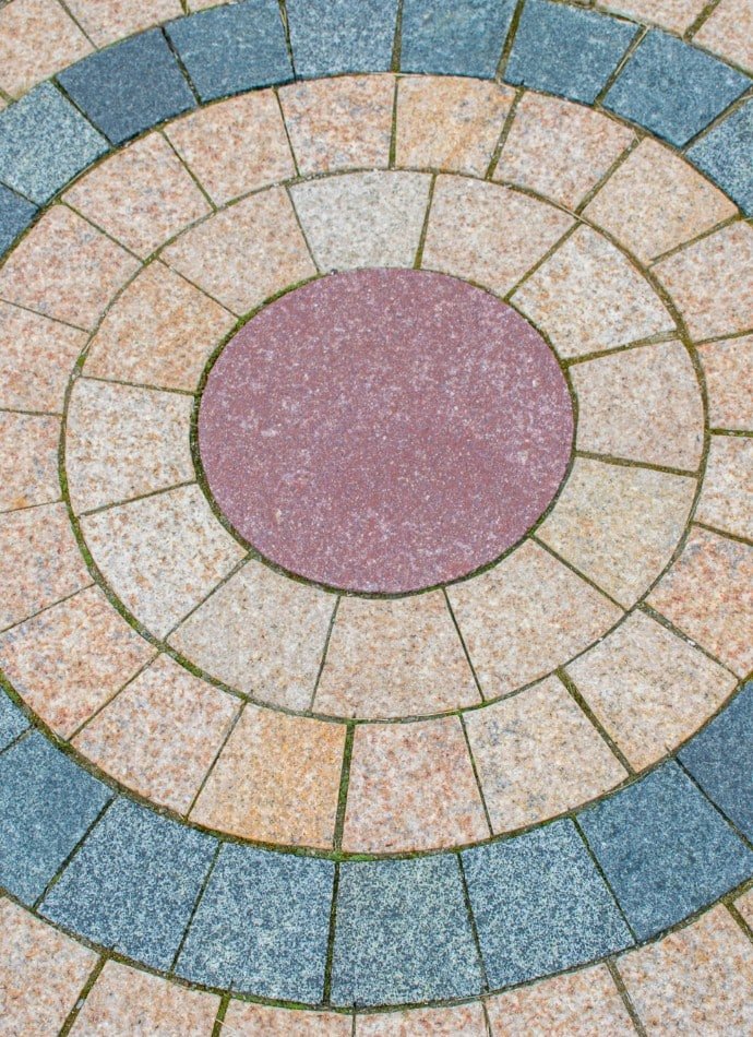 Circular patio design featuring concrete pavers in shades of red, tan, and gray. The pattern creates concentric rings, adding visual interest to the outdoor space.