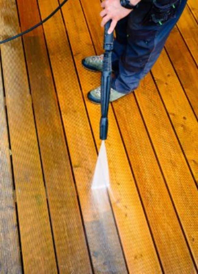 Close-up of a person power washing a wooden deck. The pressure washer blasts away dirt, revealing the clean wood grain underneath, showcasing effective fence and deck cleaning.