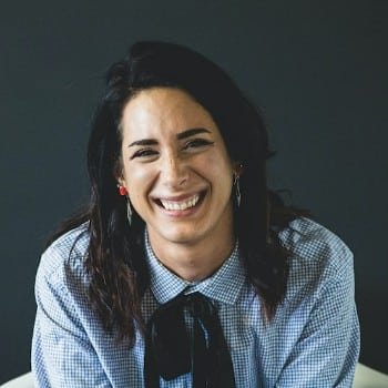 Smiling woman with dark hair wearing a blue patterned button-down shirt with a black tie. She has fair skin, dark eyebrows, and is wearing red earrings.