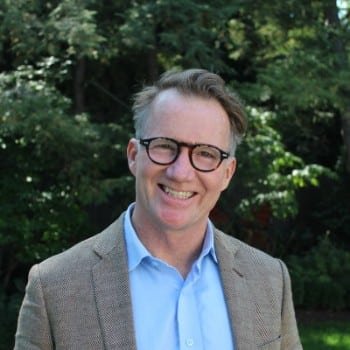 Portrait of a smiling man with glasses, wearing a blue shirt and tan blazer, standing outdoors with green trees in the background.