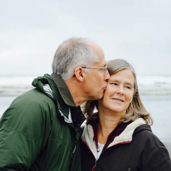 Affectionate older couple on a beach; the man kisses the smiling woman's cheek. Both wear jackets, with ocean waves visible in the background.