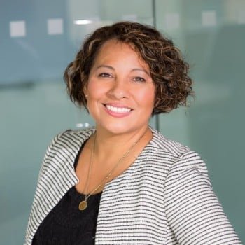 Smiling businesswoman with curly brown hair, wearing a striped blazer and gold necklace, stands confidently in front of a modern office background.