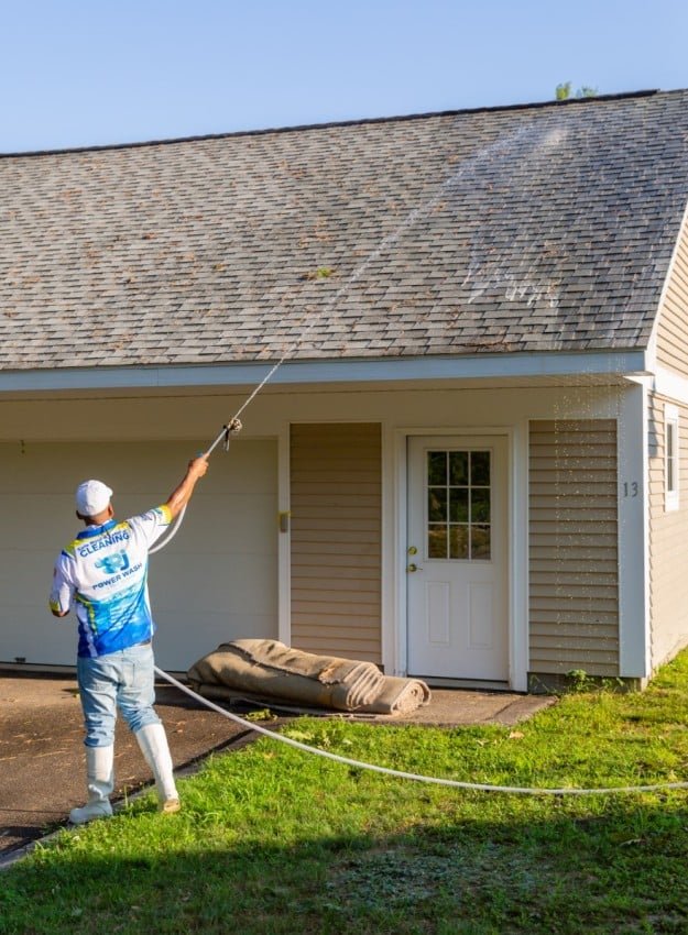 Man power washing a gray shingle roof with a long wand. A tan house with white trim is behind him. "Cleaning" and "Power Wash" are visible on his shirt.
