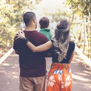 Family of three walks down a tree-lined path. The mother has her arm around the father, who is holding their child. The image conveys a sense of unity and nature.