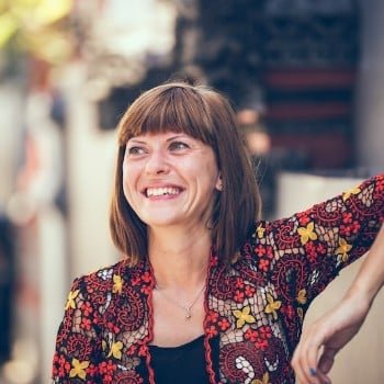 Smiling woman with bangs and a colorful floral jacket. She's outdoors, leaning against a wall, looking up and to the left.