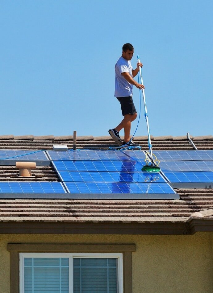 Man cleaning solar panels on a residential roof with a long-handled brush and hose. Clear blue sky above, showcasing solar panel cleaning.