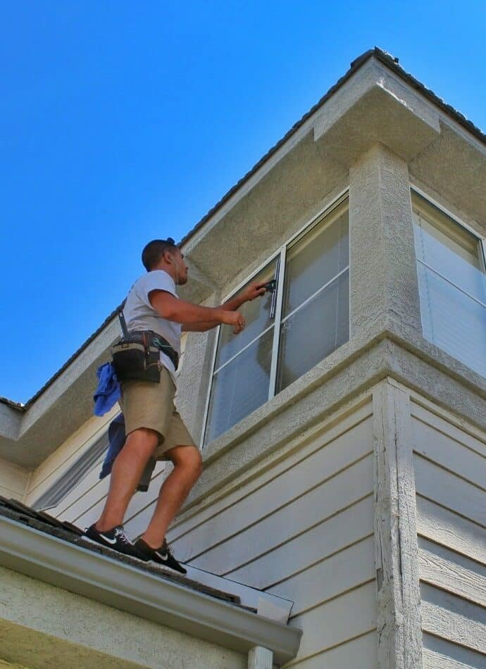 A window cleaner is shown from a low angle, standing on a roof while cleaning a second-story window. He is wearing a tool belt and using a squeegee to clean the window. Blue skies in the background.