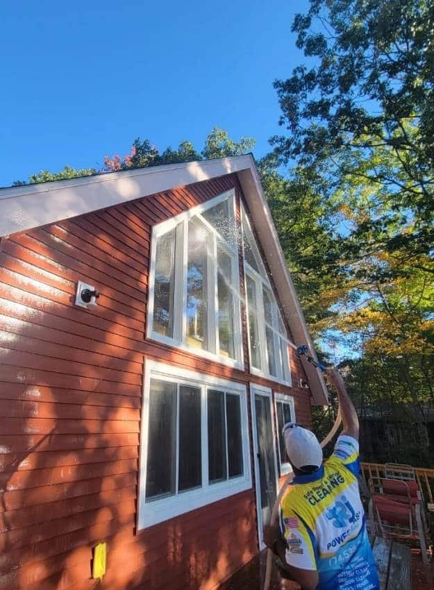 Man power washing windows of a red house with white trim under a blue sky. "Get Busy Cleaning" is printed on his shirt. Window cleaning in progress.