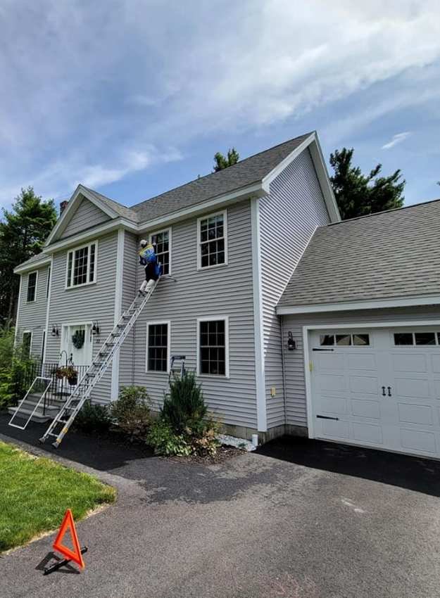 A professional window cleaner on a ladder washes the windows of a gray two-story house with a gray roof. An orange safety cone sits on the driveway.