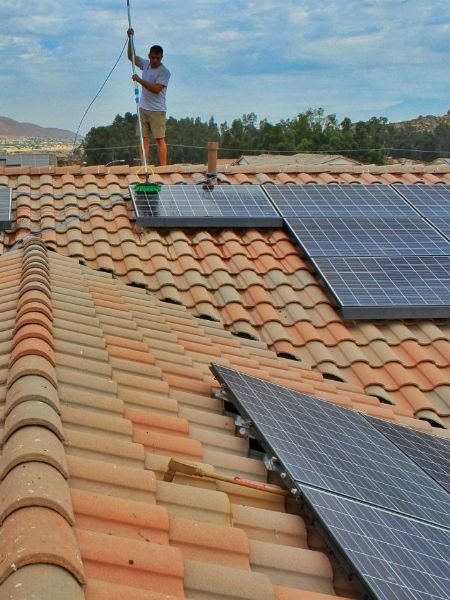 Man cleaning solar panels on a terracotta tile roof with a long-handled brush. Residential solar panel cleaning helps maintain efficiency.
