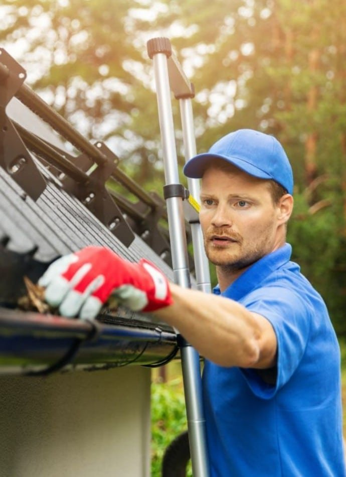 Man on ladder cleaning gutters, wearing blue shirt, hat, and red gloves. Focus on gutter cleaning maintenance as part of home upkeep.