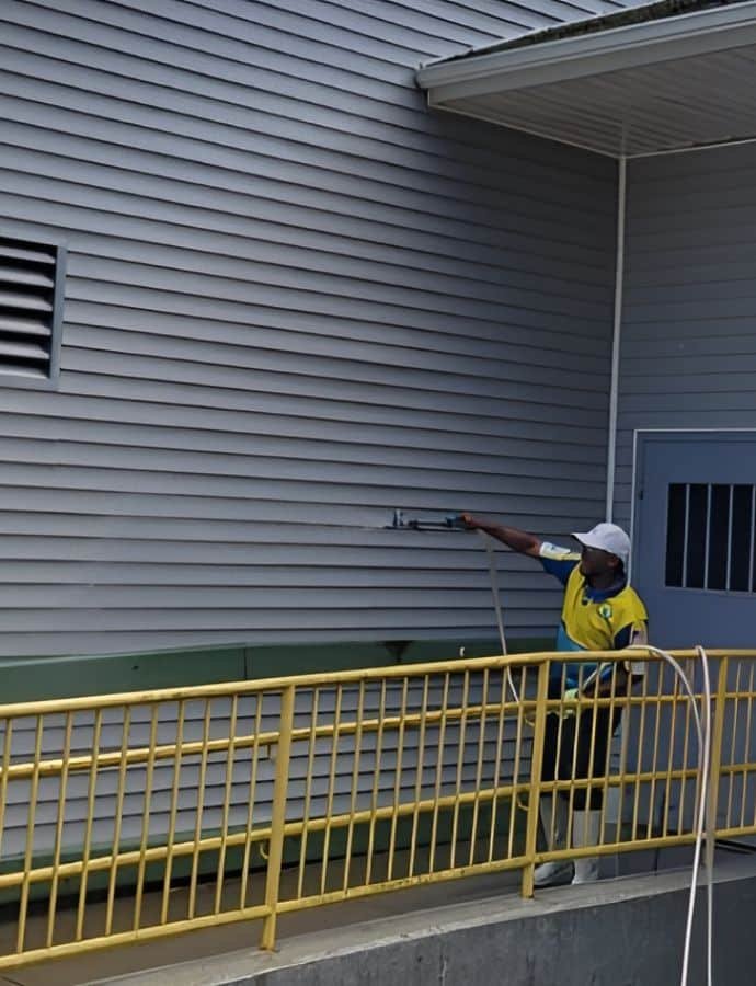 A worker performs commercial power washing on a gray building exterior. He uses a pressure washer wand while standing on a yellow-railed walkway.