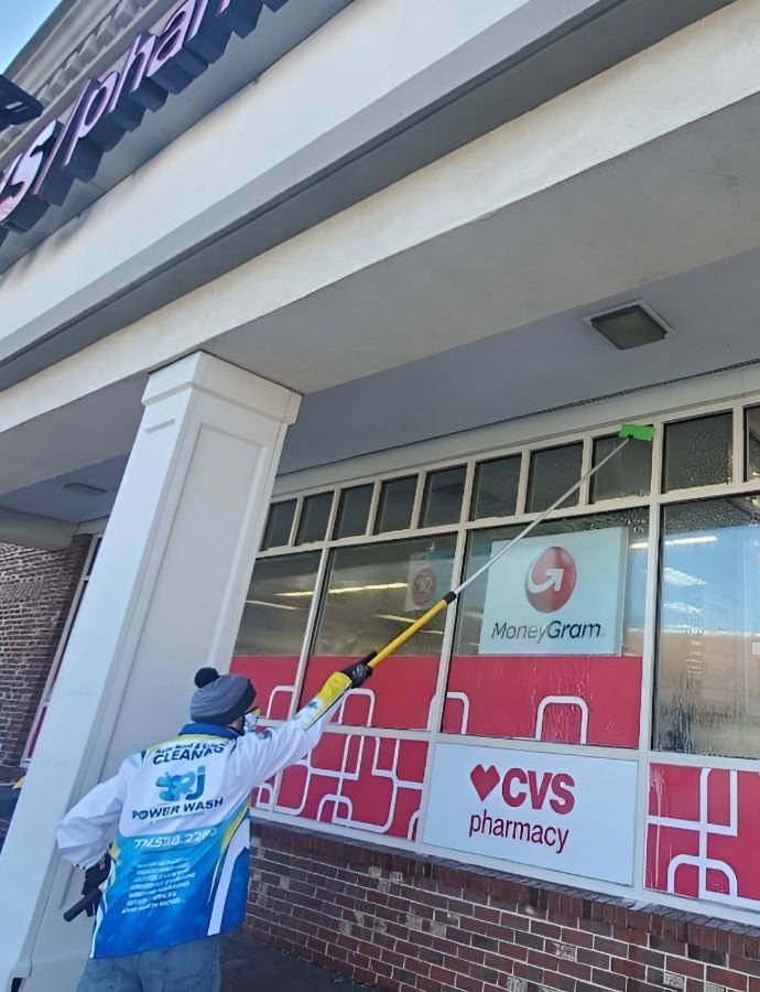 A worker uses a long pole to power wash the windows of a CVS pharmacy. The building's facade is brick and the windows have MoneyGram and CVS Pharmacy signage.